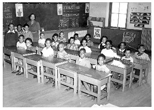 Black and white photograph of a third grade class, African American students seated at desks looking at the photographer, their teacher standing at a blackboard behind them.><p style=