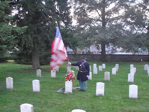 A lone African American color bearer in a Union Civil War uniform stands amidst rows of headstones at Gettysburg National Cemetery.